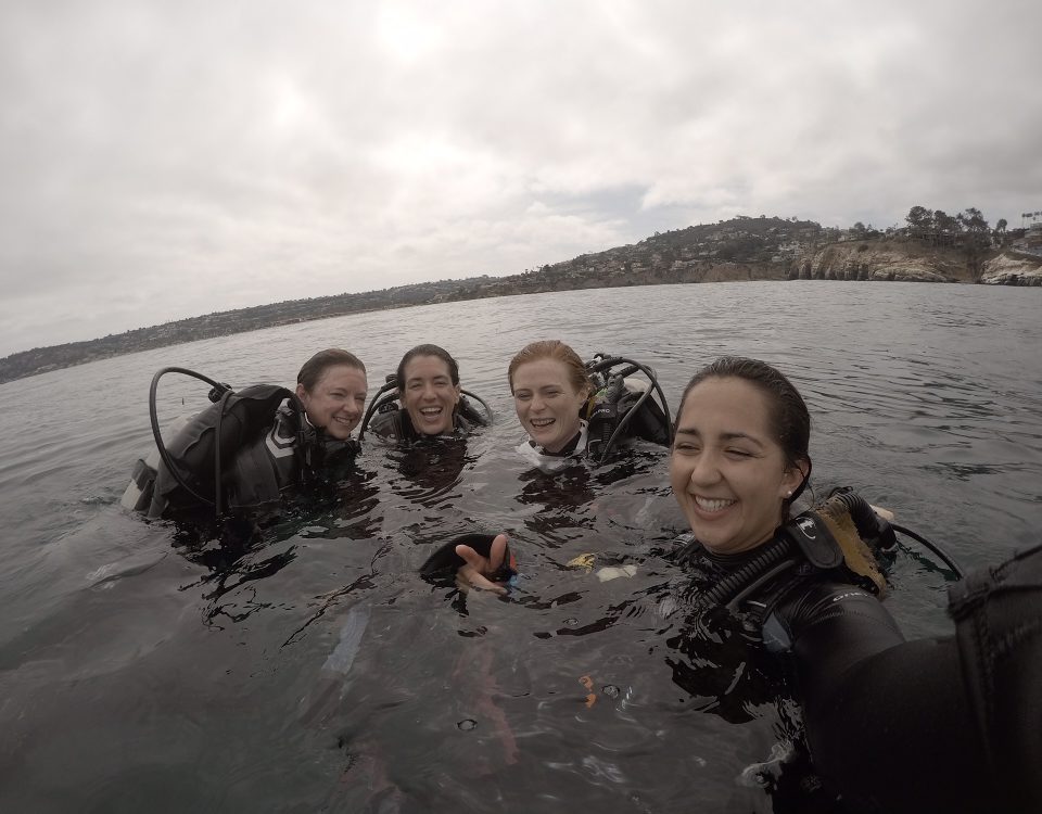 Four women in scuba gear, at the surface of the water smiling and laughing